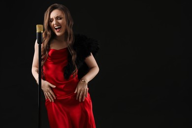 Beautiful young woman in stylish dress with microphone singing on black background, space for text