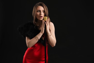 Beautiful young woman in stylish dress with microphone singing on black background, space for text