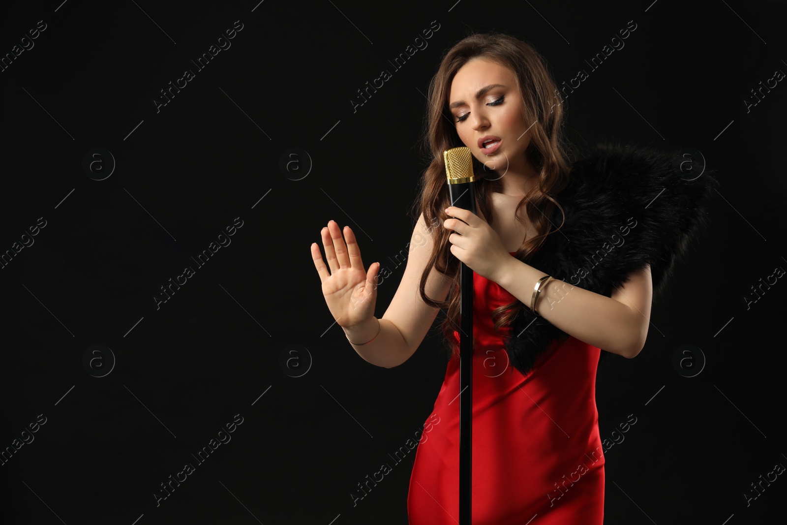 Photo of Beautiful young woman in stylish dress with microphone singing on black background, space for text