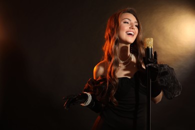 Photo of Beautiful young woman in stylish dress with microphone singing on dark background, space for text