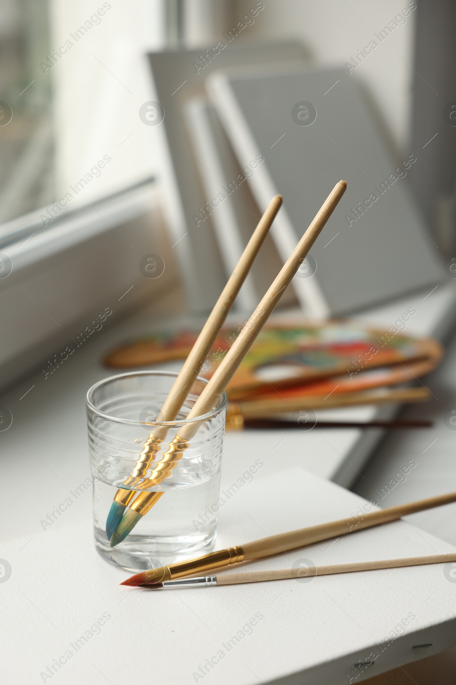 Photo of Brushes, glass of water and blank canvases on window sill, closeup