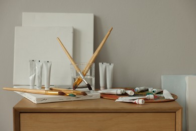 Photo of Artist's palette, brushes, canvases and paints on wooden chest of drawers indoors