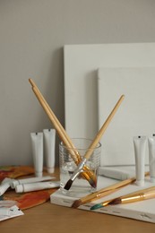 Photo of Artist's palette, brushes in glass of water, blank canvases and paints on wooden table indoors