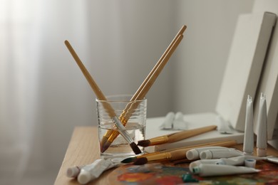 Artist's palette, brushes in glass of water and paints on wooden table indoors