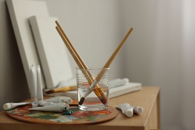 Photo of Artist's palette, brushes in glass of water and paints on wooden table indoors