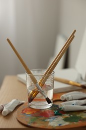 Photo of Artist's palette, brushes in glass of water and paints on wooden table indoors