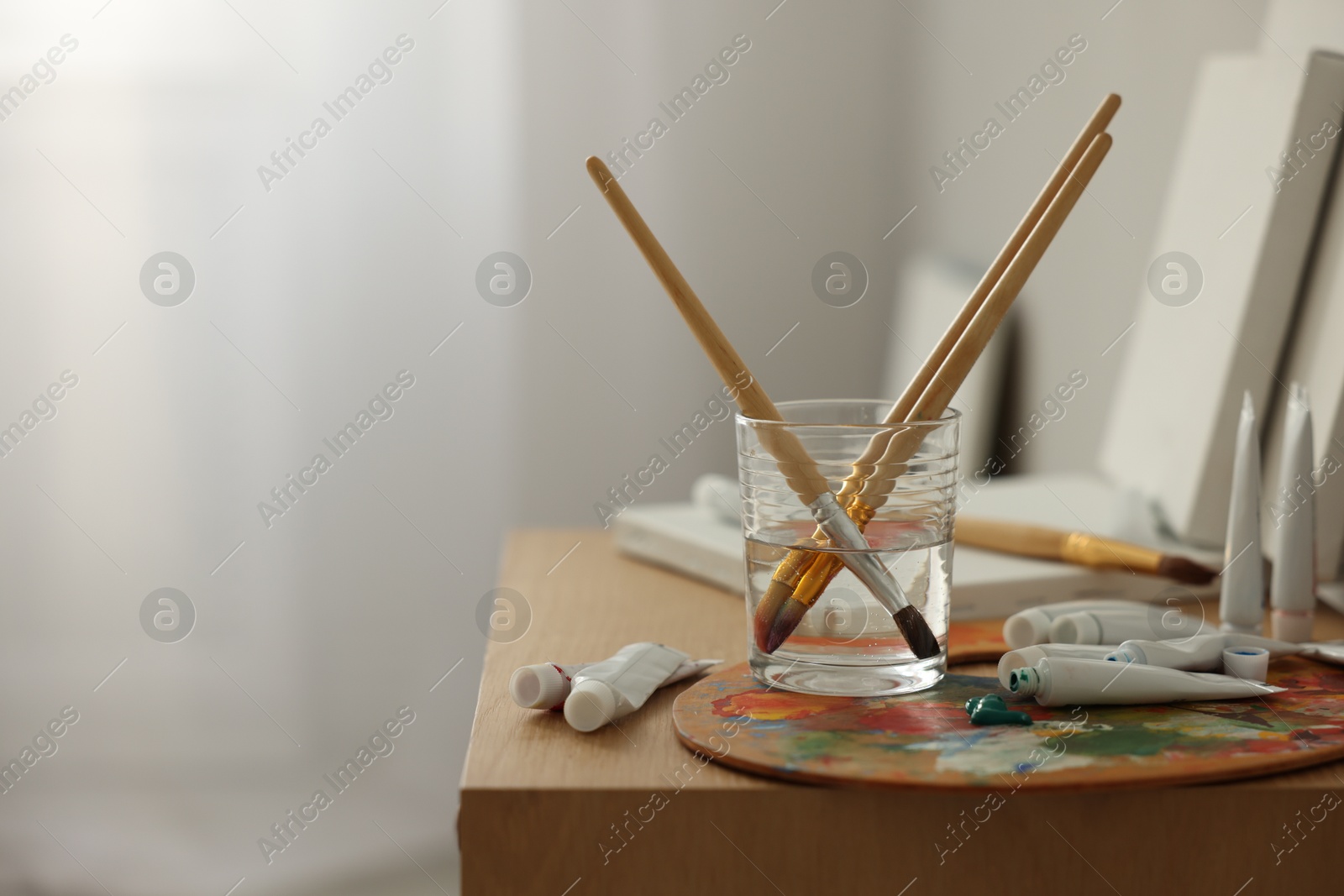 Photo of Artist's palette, brushes in glass of water and paints on wooden table indoors. Space for text