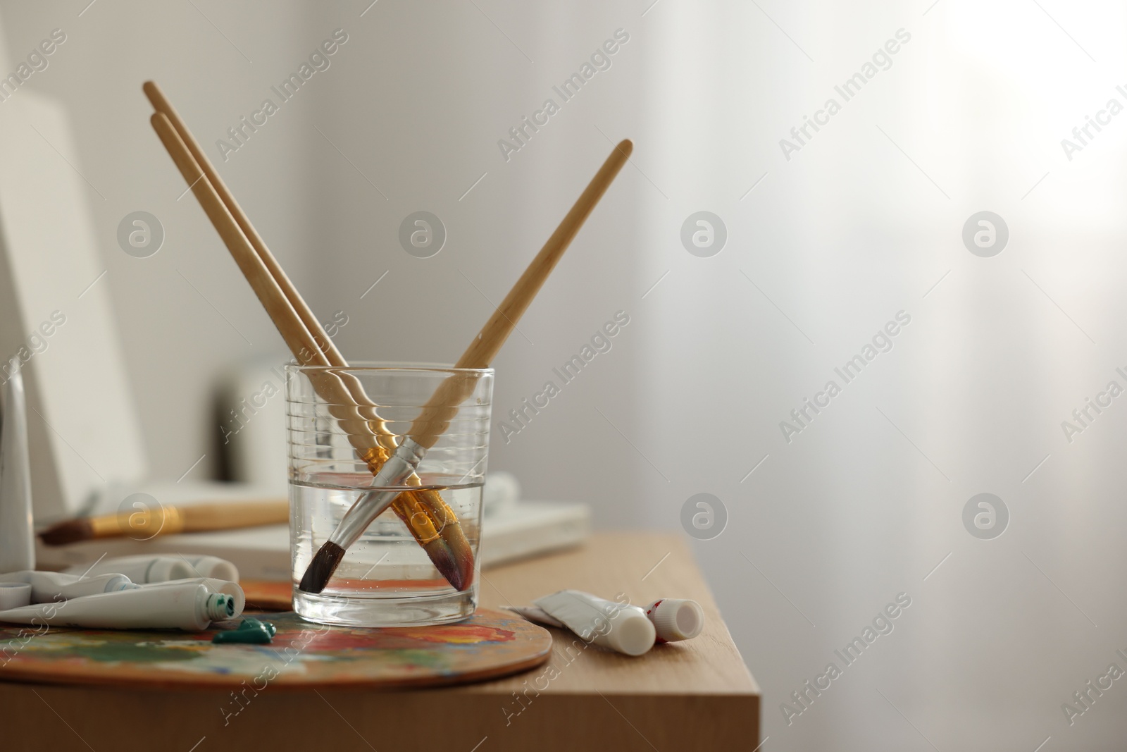 Photo of Artist's palette, brushes in glass of water and paints on wooden table indoors. Space for text