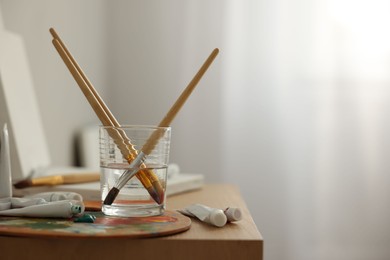 Artist's palette, brushes in glass of water and paints on wooden table indoors. Space for text