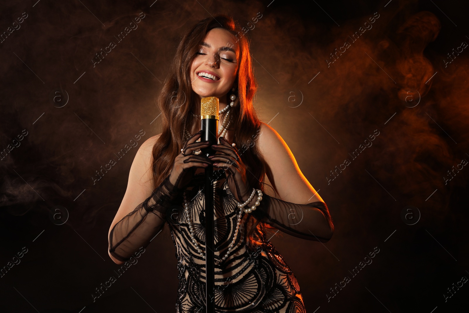 Photo of Beautiful young woman in stylish dress with microphone singing on dark background with smoke