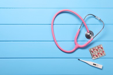 Photo of Stethoscope, thermometer and pills on light blue wooden background, flat lay. Space for text