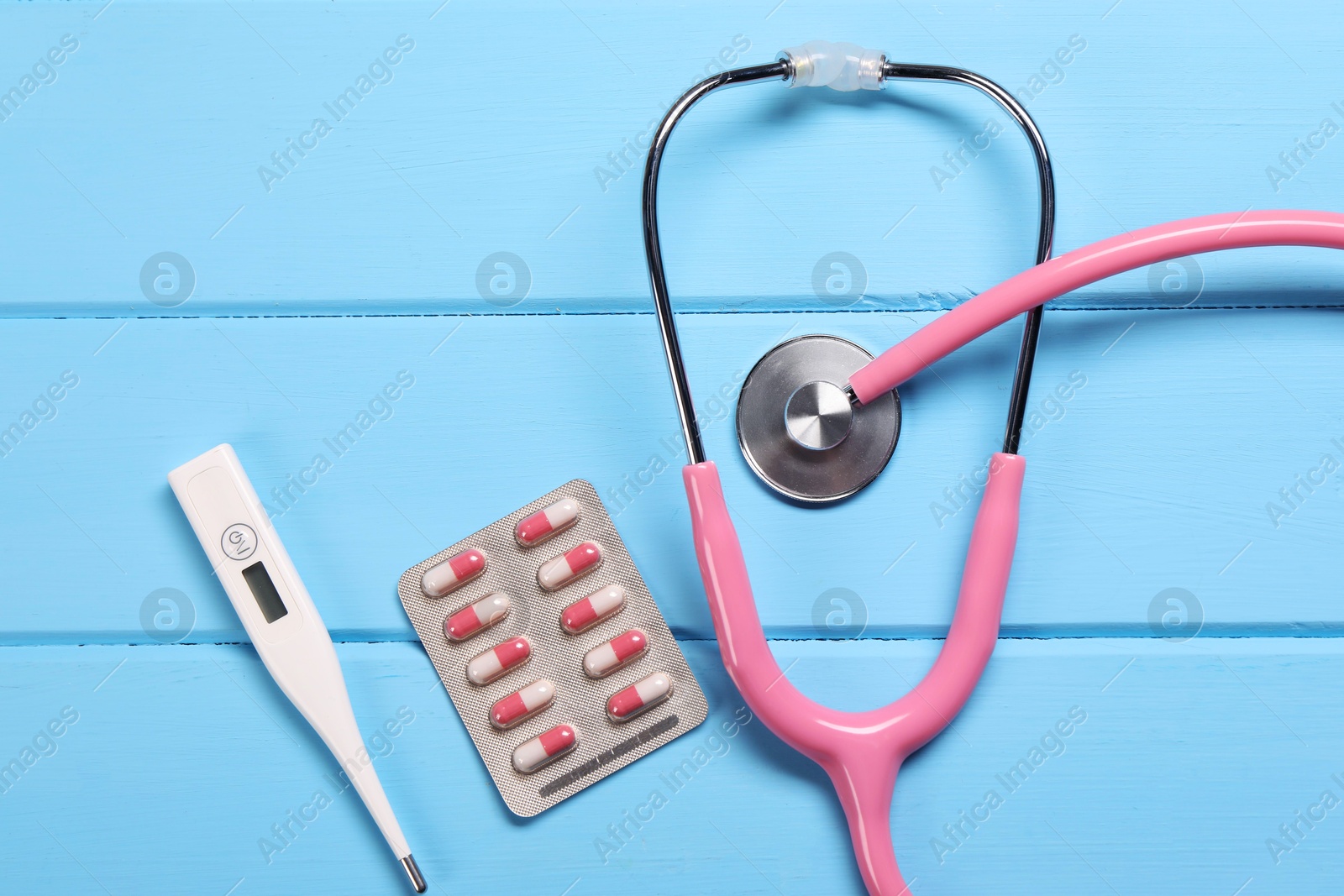 Photo of Stethoscope, thermometer and pills on light blue wooden background, flat lay. Medical treatment