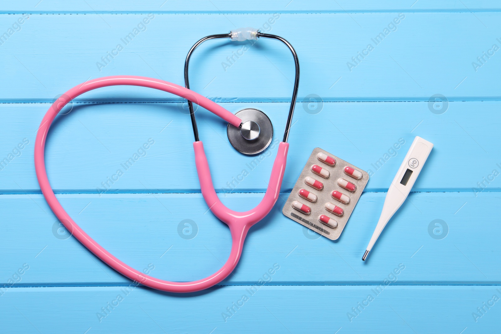 Photo of Stethoscope, thermometer and pills on light blue wooden background, flat lay. Medical treatment