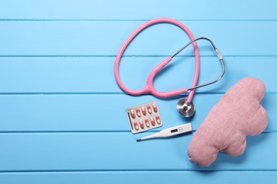 Pillow, stethoscope, thermometer and pills on light blue wooden background, flat lay. Space for text