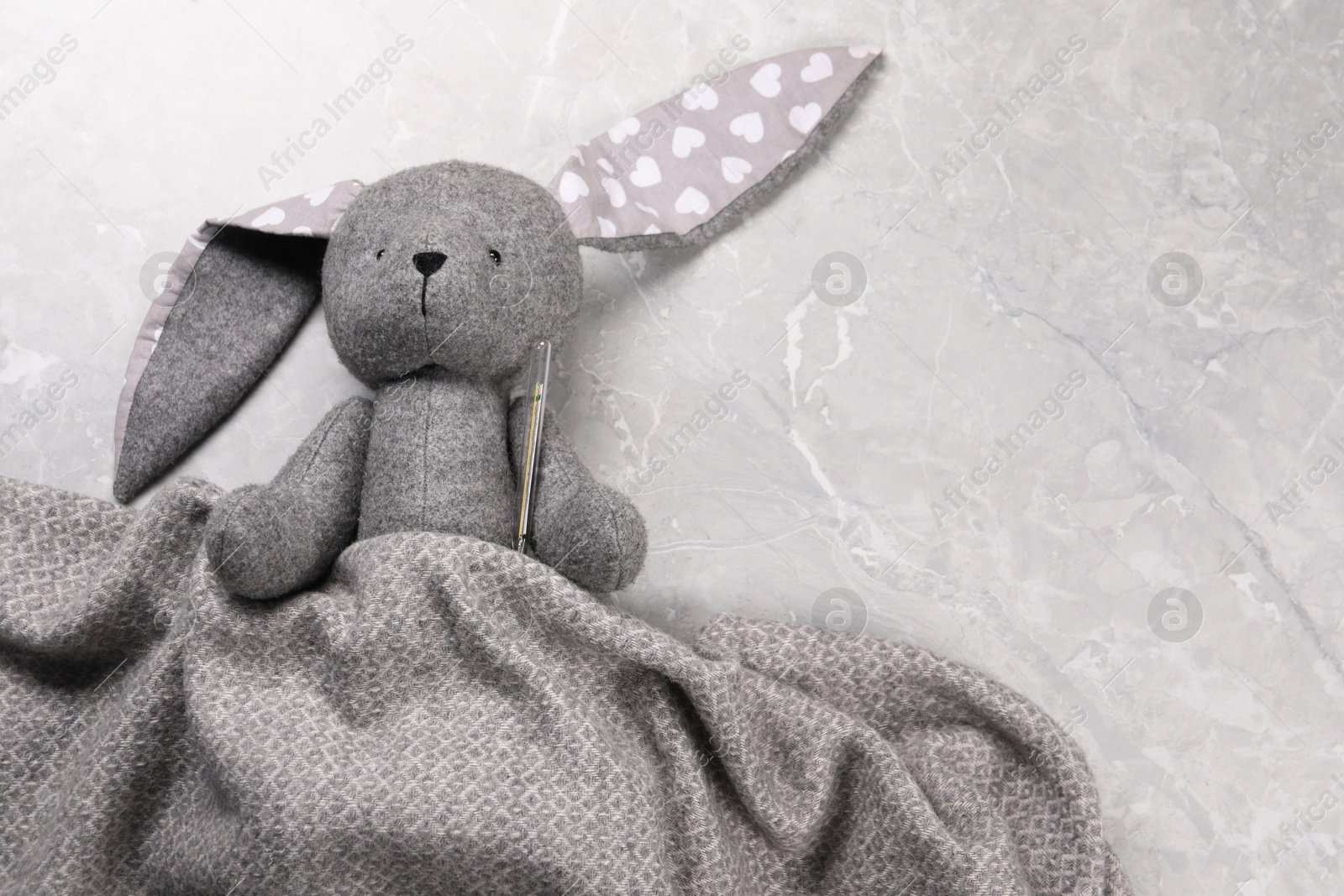 Photo of Toy bunny with thermometer under blanket on gray marble background, top view. Space for text
