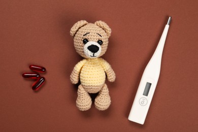 Toy bear, thermometer and pills on brown background, flat lay