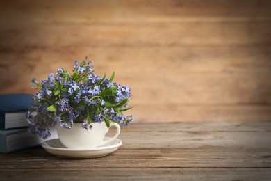 Photo of Beautiful forget-me-not flowers in cup, saucer and books on wooden table. Space for text