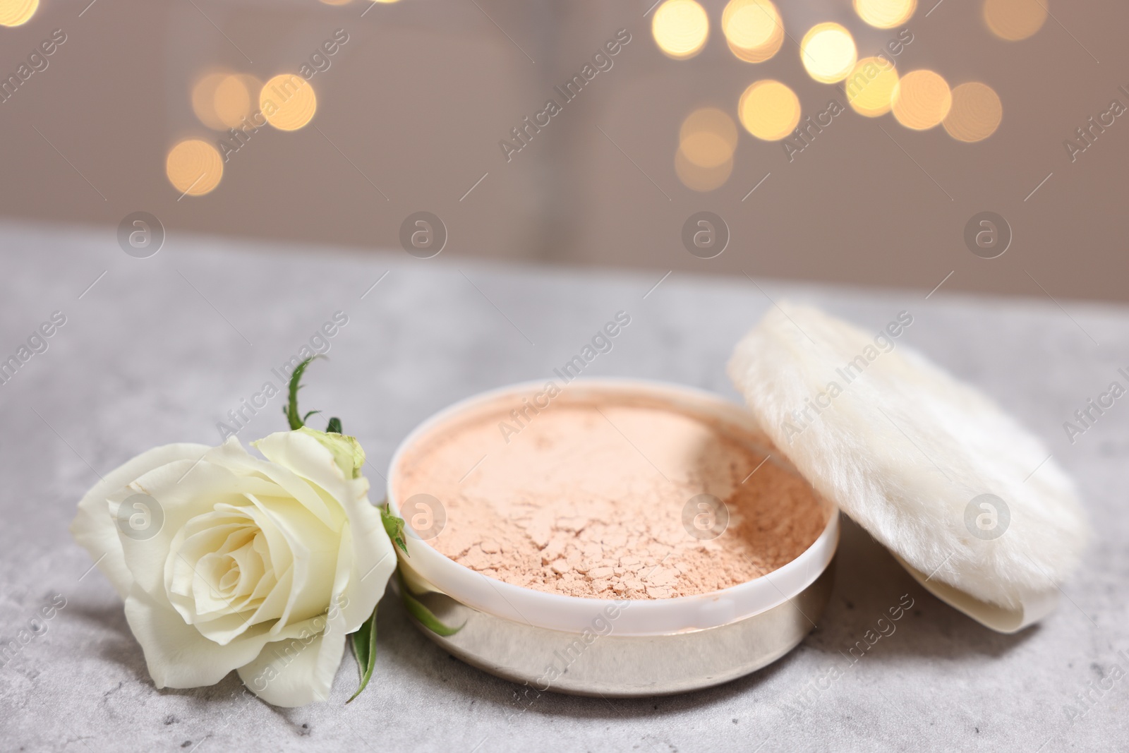Photo of Face powder, puff applicator and rose flower on grey textured table against blurred lights, closeup