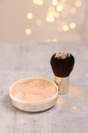 Face powder and brush on grey textured table against blurred lights, closeup