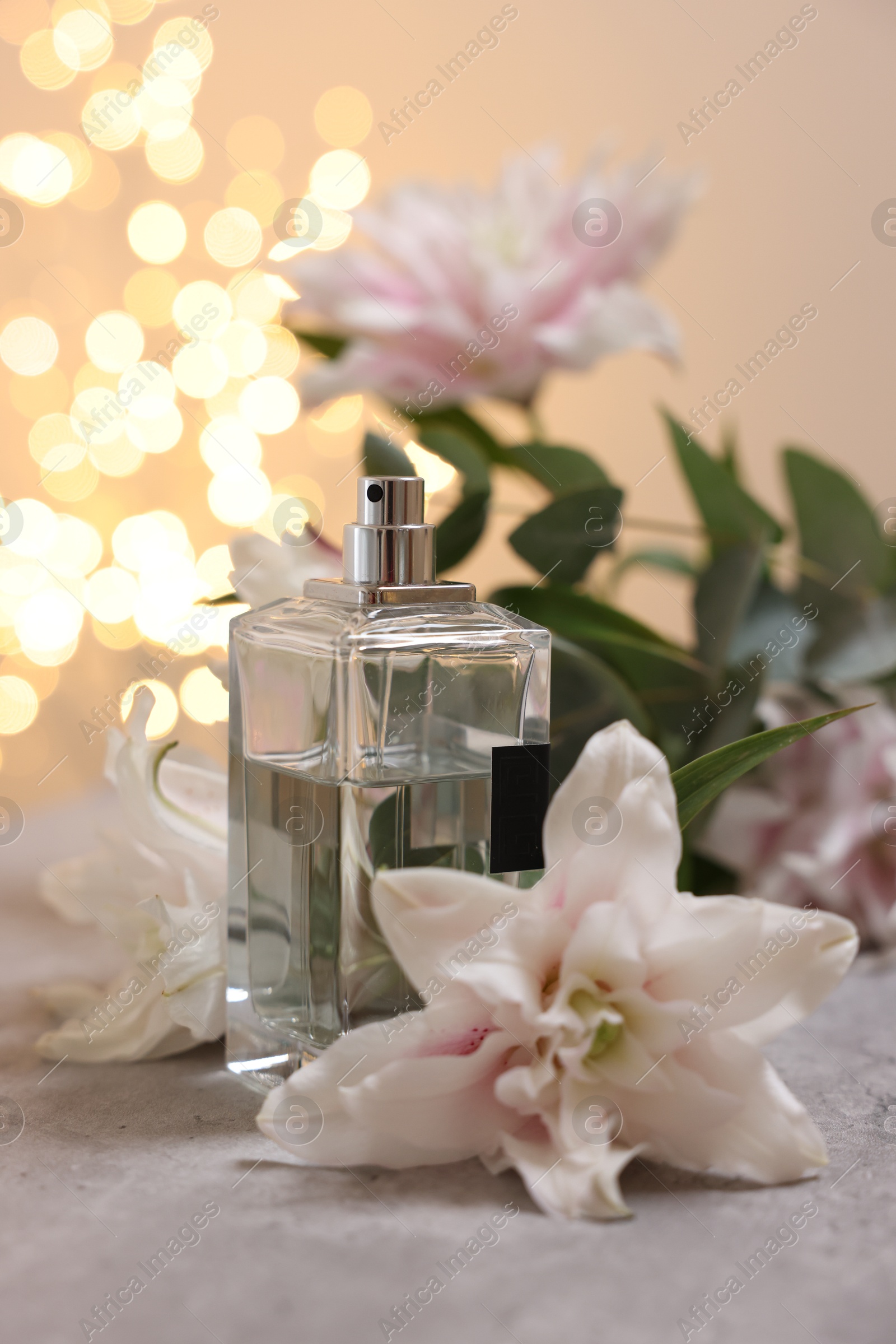 Photo of Bottle of perfume and beautiful lily flowers on table against beige background with blurred lights, closeup