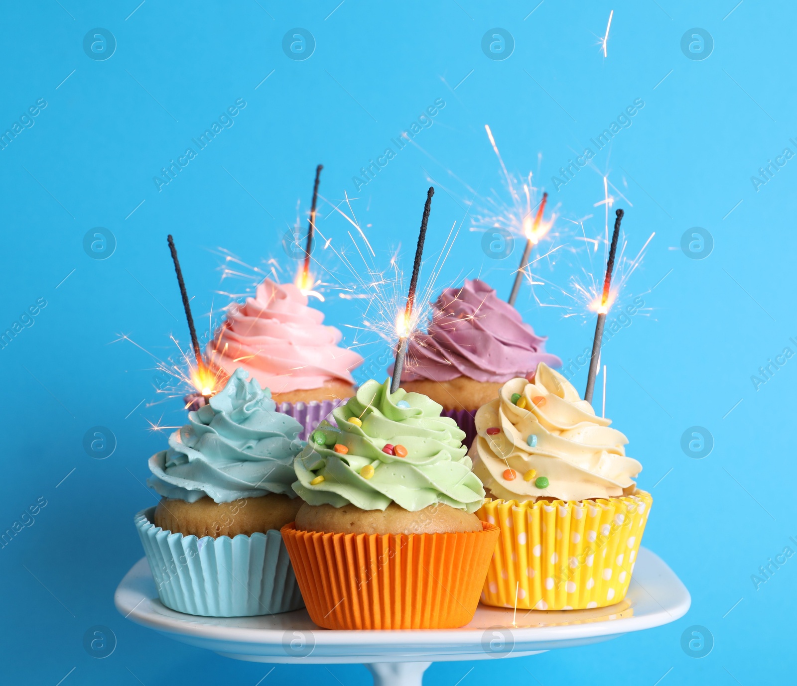 Photo of Birthday cupcakes with burning sparklers on stand against light blue background