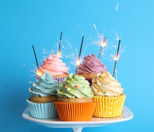 Birthday cupcakes with burning sparklers on stand against light blue background