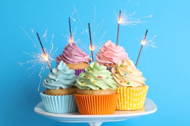 Photo of Birthday cupcakes with burning sparklers on stand against light blue background