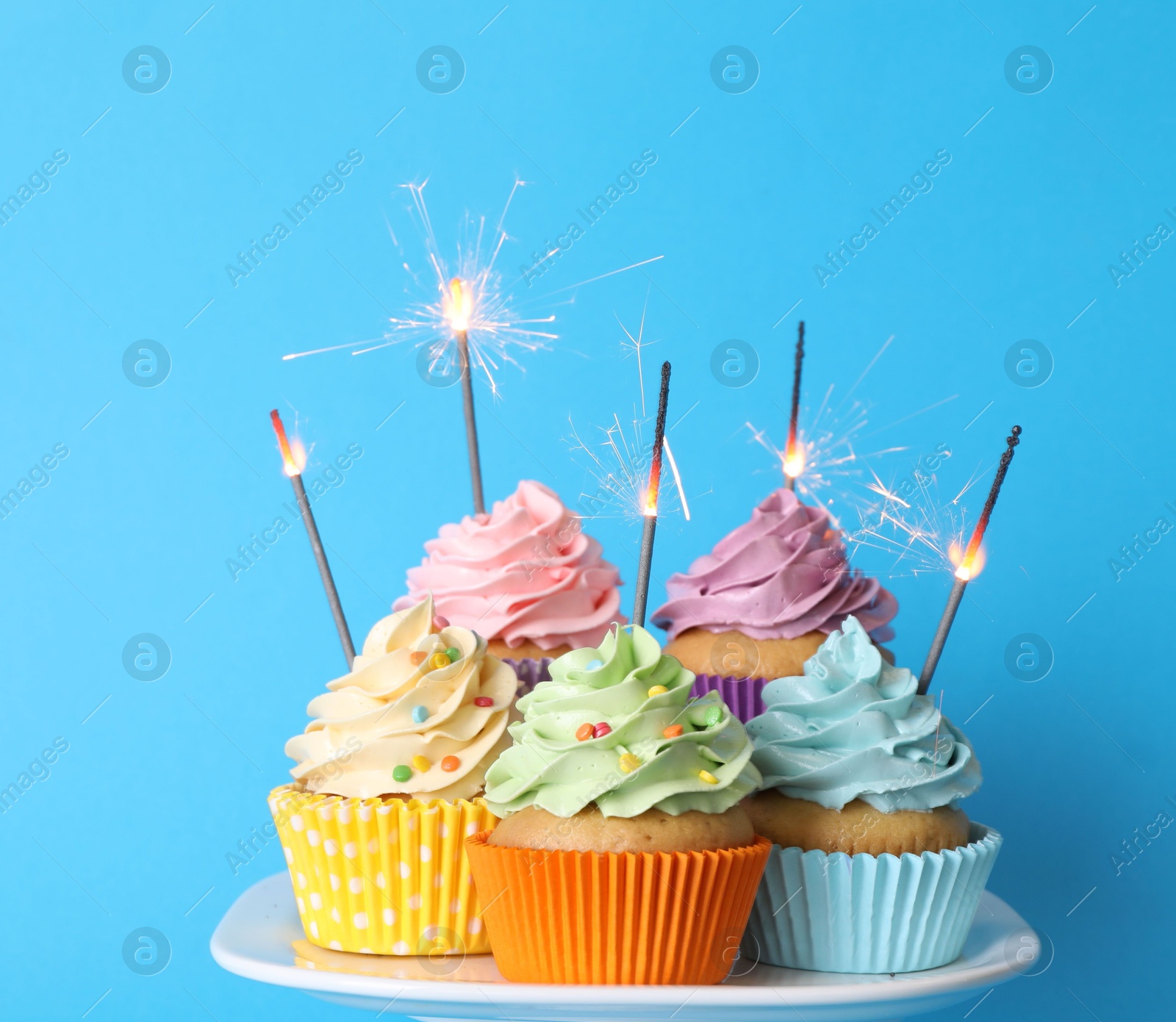 Photo of Birthday cupcakes with burning sparklers on stand against light blue background