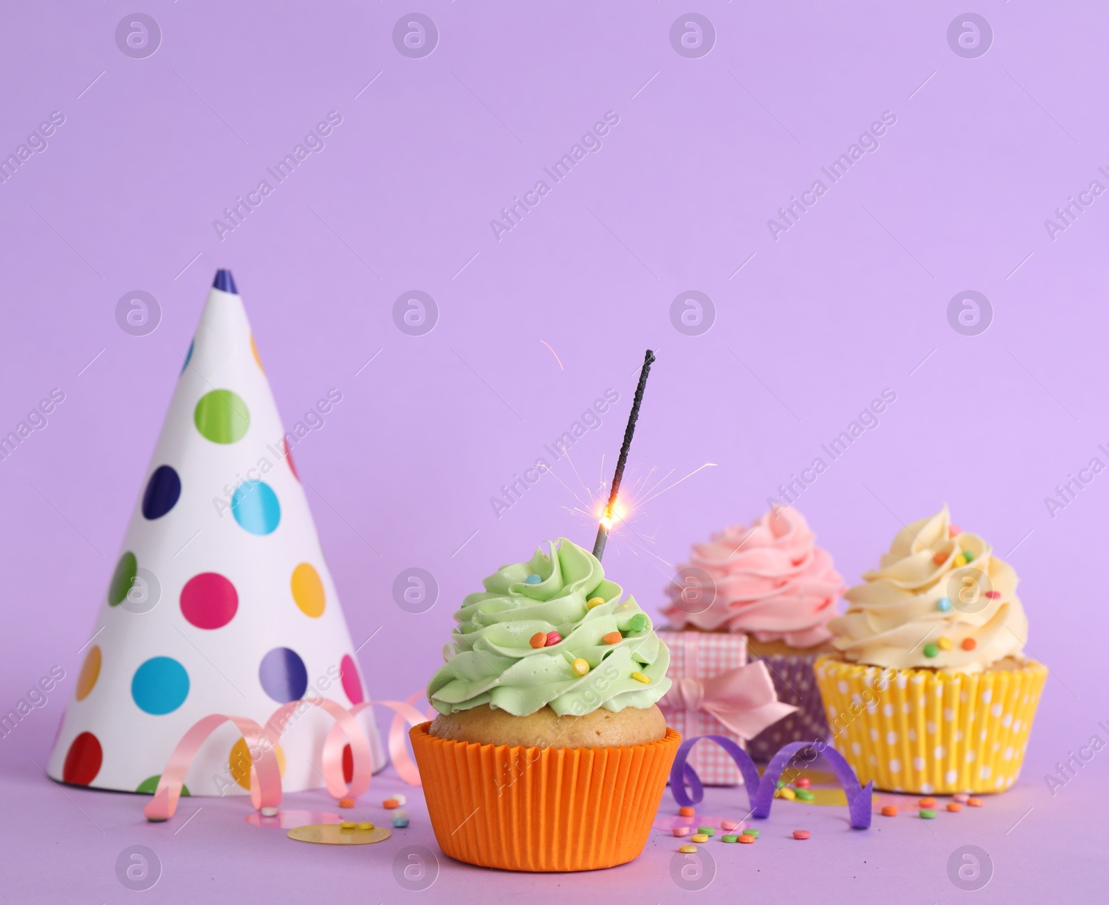 Photo of Birthday cupcakes with burning sparkler, party hat and gift box on violet background