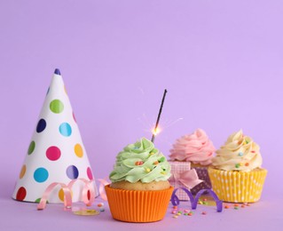 Photo of Birthday cupcakes with burning sparkler, party hat and gift box on violet background