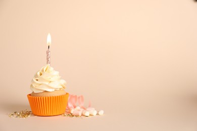 Delicious birthday cupcake with burning candle, marshmallows and streamer on beige background, space for text