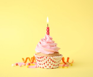 Photo of Delicious birthday cupcake with burning candle, sprinkles and streamer on yellow background