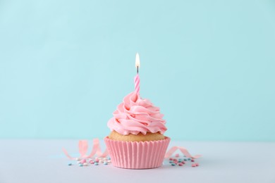 Delicious birthday cupcake with burning candle, sprinkles and streamer on white table against turquoise background