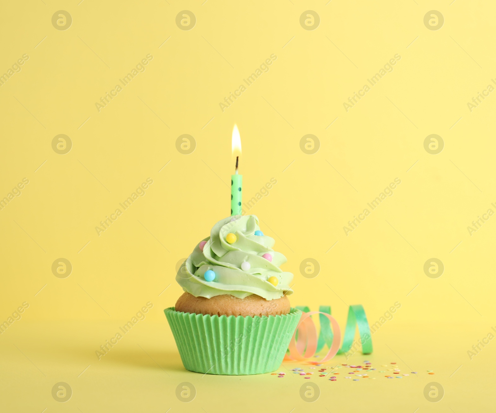 Photo of Delicious birthday cupcake with burning candle, sprinkles and streamers on yellow background