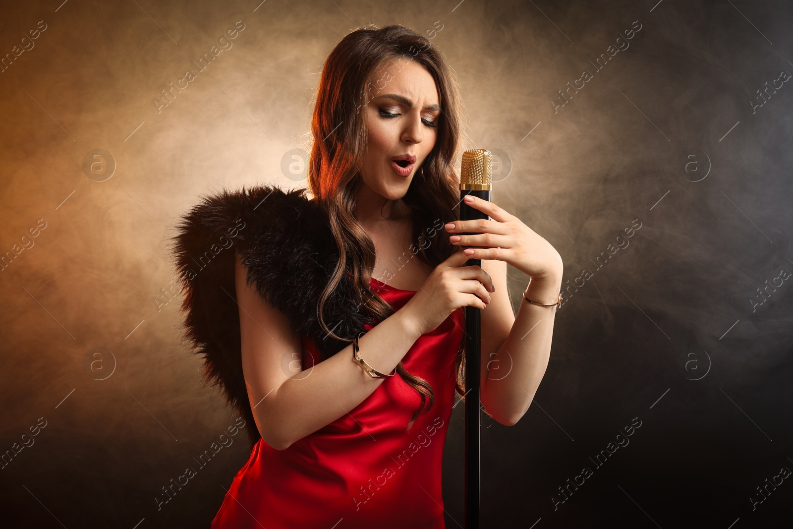 Photo of Beautiful young woman in stylish dress with microphone singing on dark background in color lights and smoke