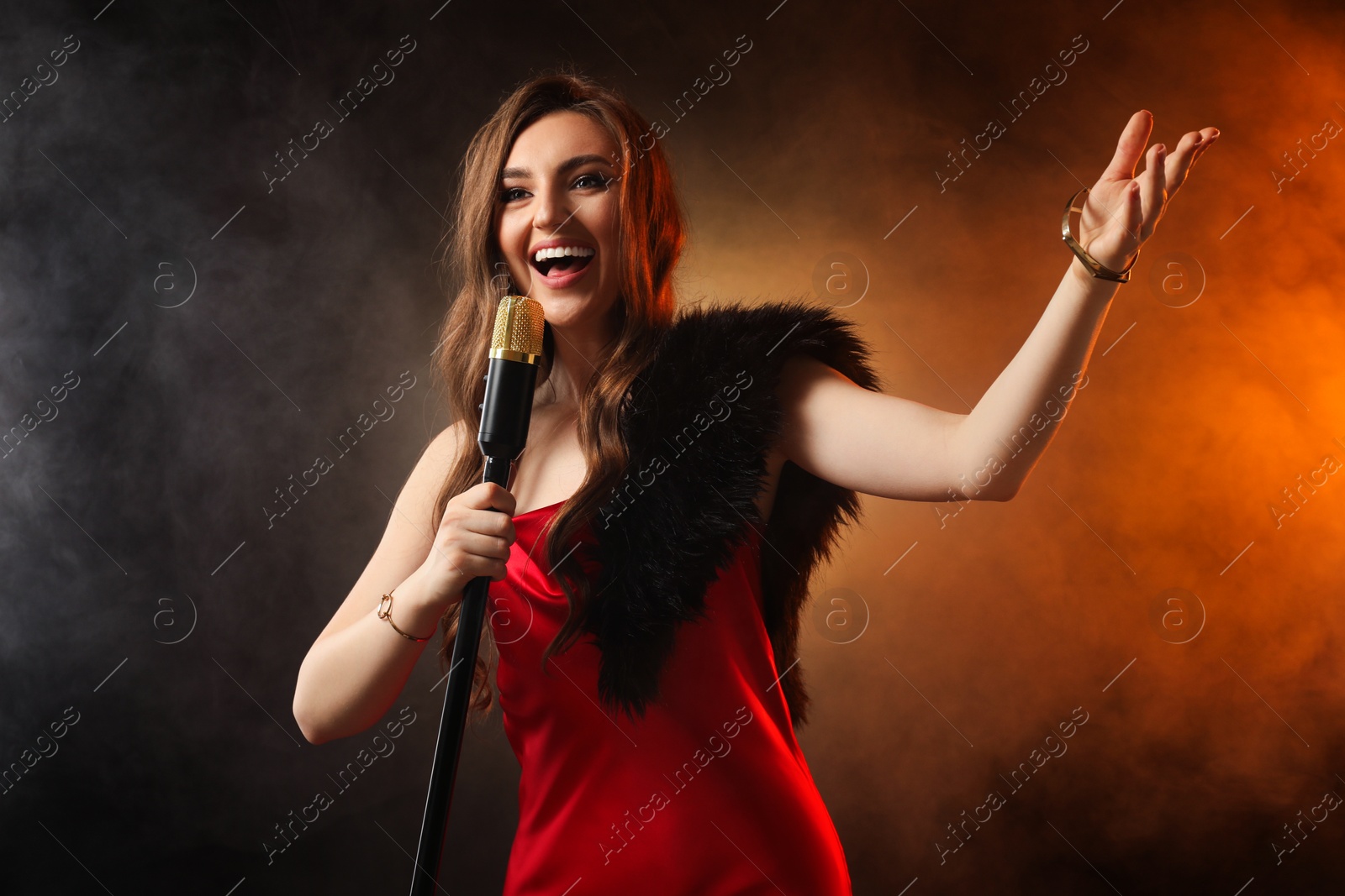 Photo of Beautiful young woman in stylish dress with microphone singing on dark background in color lights and smoke