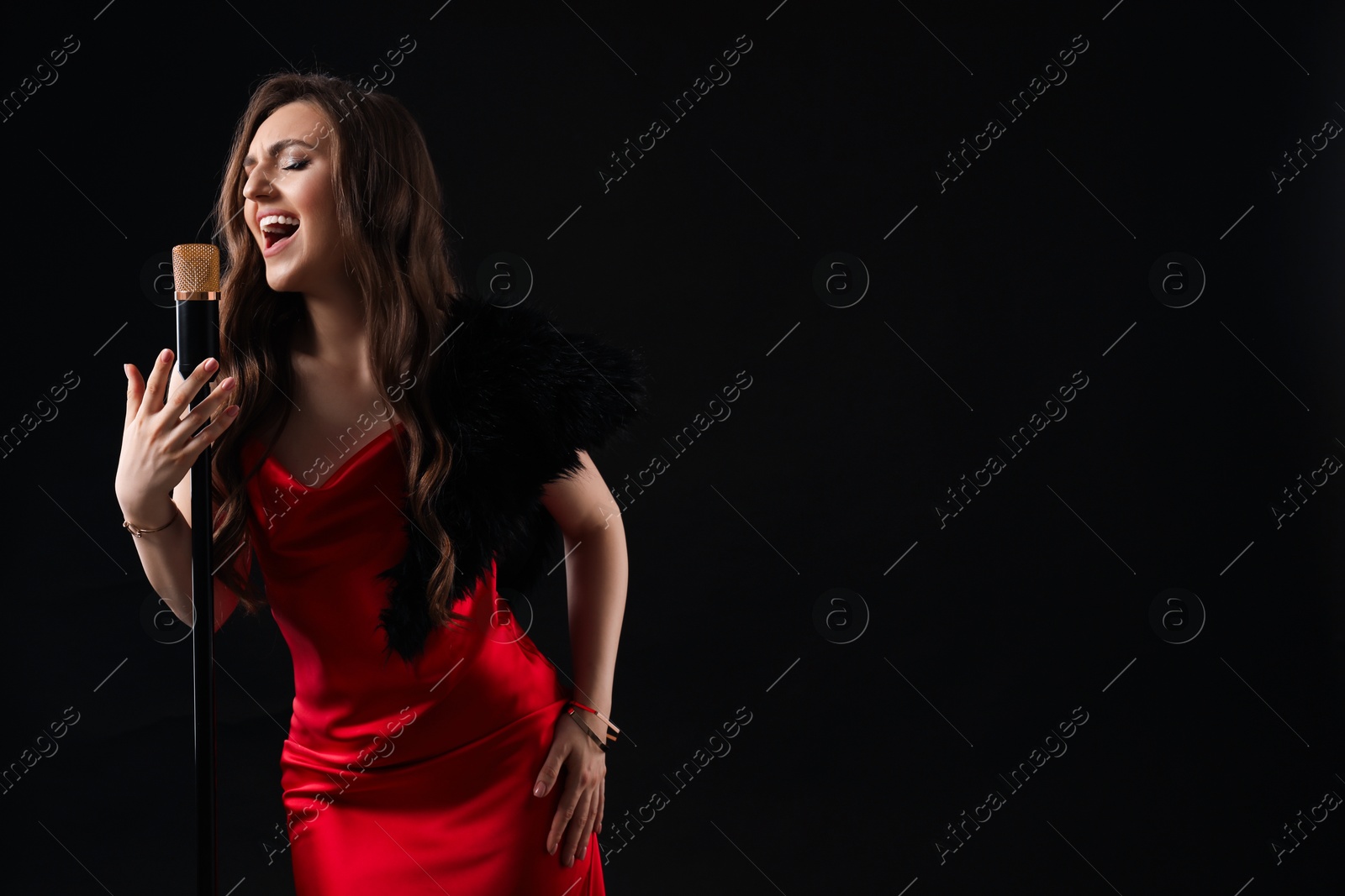 Photo of Beautiful young woman in stylish dress with microphone singing on black background, space for text