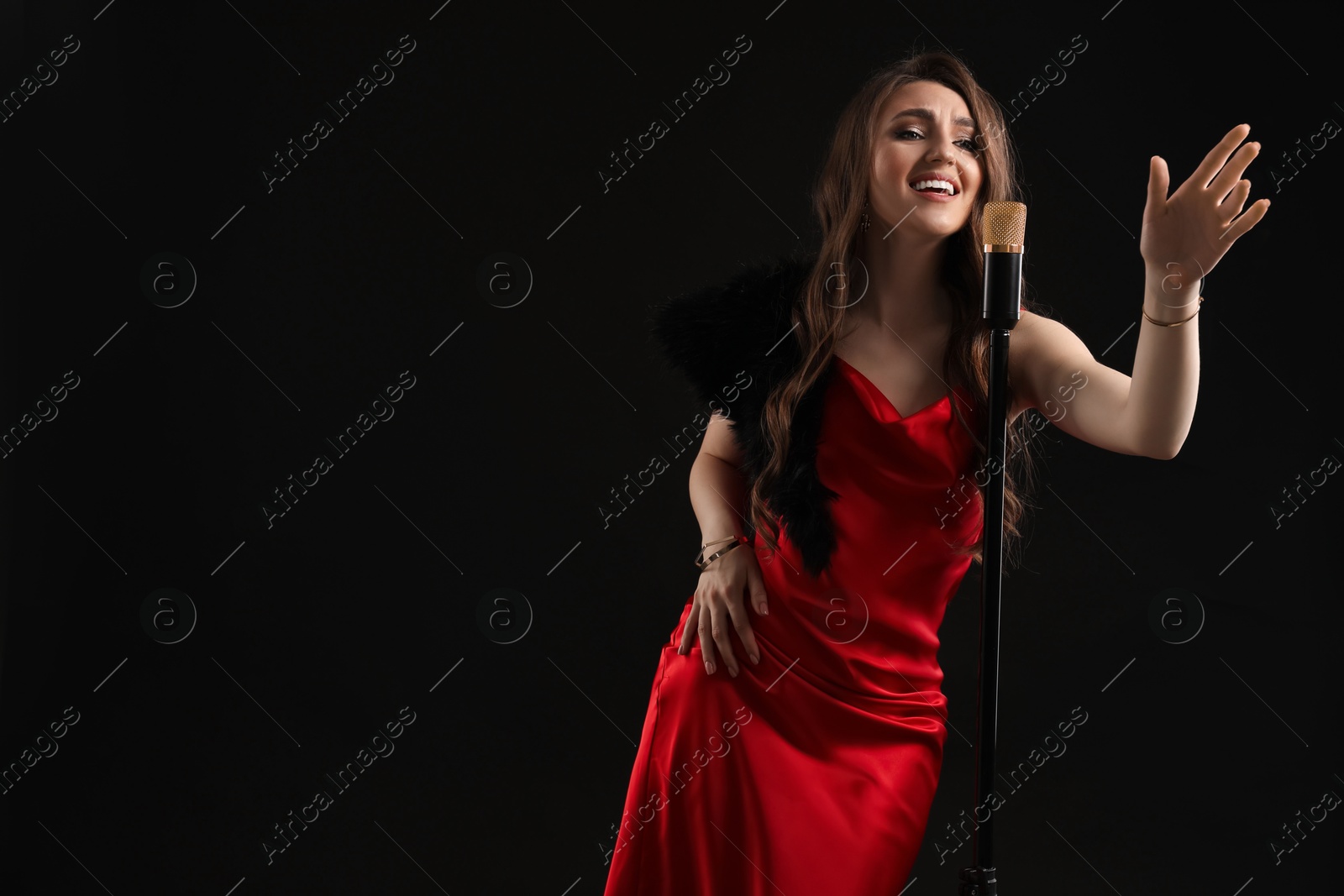 Photo of Beautiful young woman in stylish dress with microphone singing on black background, space for text