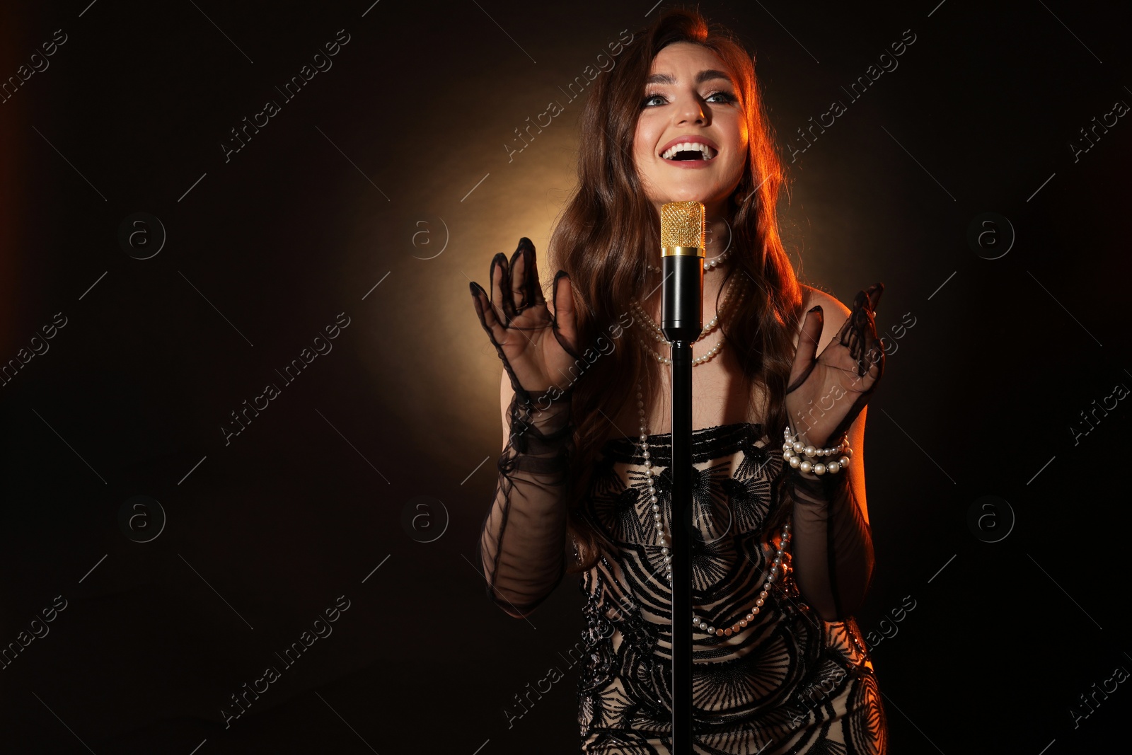 Photo of Beautiful young woman in stylish dress with microphone singing on dark background, space for text