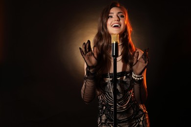 Beautiful young woman in stylish dress with microphone singing on dark background, space for text