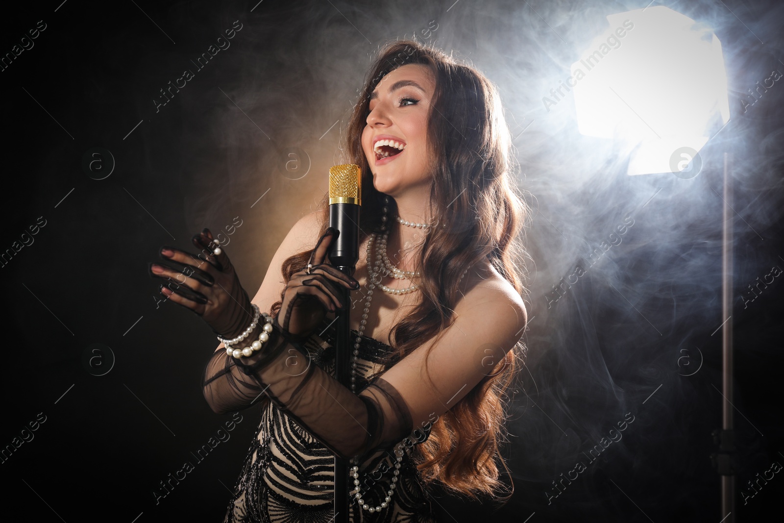 Photo of Beautiful young woman with microphone singing on dark background with smoke