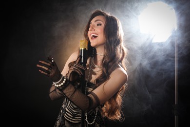 Beautiful young woman with microphone singing on dark background with smoke