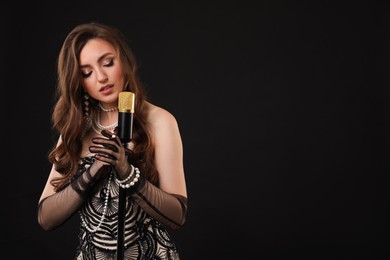 Beautiful young woman in stylish dress with microphone singing on black background, space for text