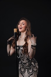 Beautiful young woman in stylish dress with microphone singing on black background