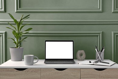 Image of Stylish workspace with laptop and houseplant on wooden desk near sage green wall