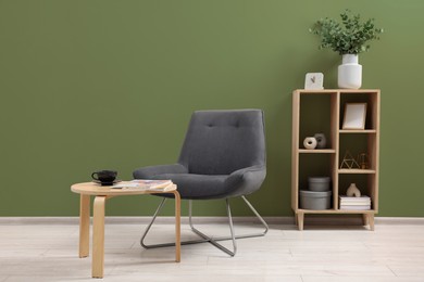 Image of Comfortable armchair, coffee table and shelving unit near sage green wall indoors. Interior design