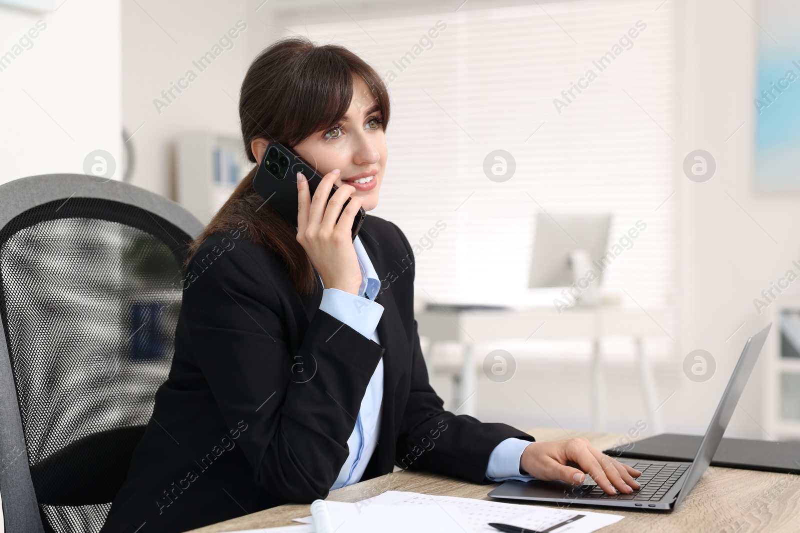 Photo of Smiling secretary talking by smartphone at table in office