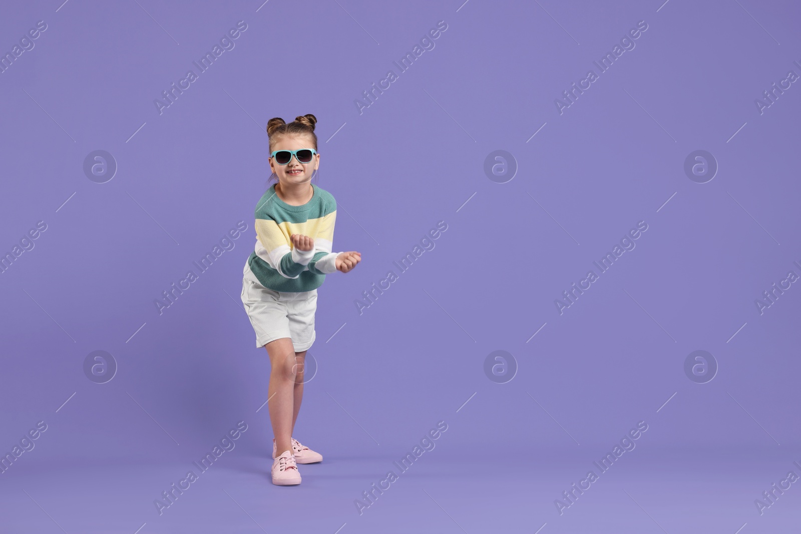 Photo of Cute little girl in sunglasses dancing on violet background, space for text