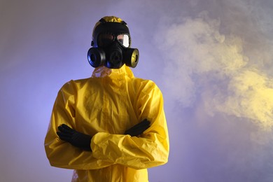 Worker wearing gas mask in smoke on violet background, low angle view. Space for text