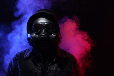 Photo of Man wearing gas mask in color lights and smoke on black background. Space for text